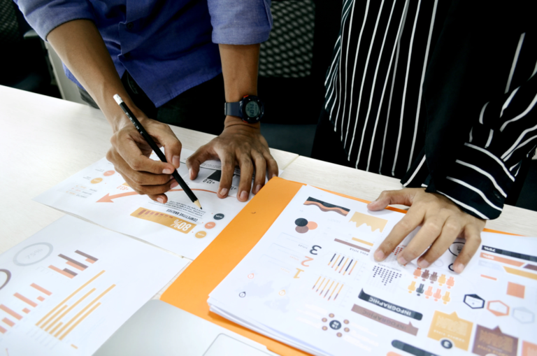 2 people stand over printouts of data, consisting of charts and graphs